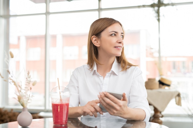 Bastante joven mujer serena desplazándose en el teléfono inteligente mientras está sentado en la cafetería, tomando una copa y eligiendo productos en la tienda online