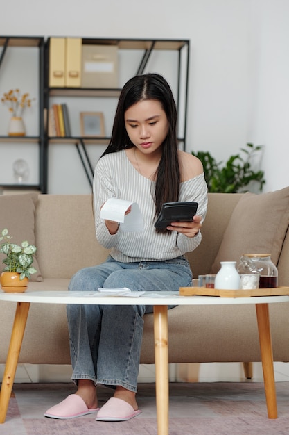 Bastante joven mujer vietnamita sentada en un sofá en la sala de estar revisando las facturas y calculando los gastos mensuales