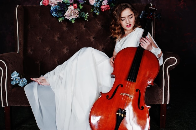 Bastante joven músico gilrl en vestido blanco con contrabajo sentado en un sofá vintage marrón