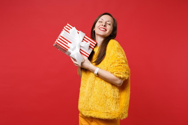 Bastante joven con los ojos cerrados mantenga el presente cuadro de rayas rojas con cinta de regalo aislada sobre fondo rojo. Día de San Valentín, Día Internacional de la Mujer, cumpleaños, concepto de vacaciones. Simulacros de espacio de copia.