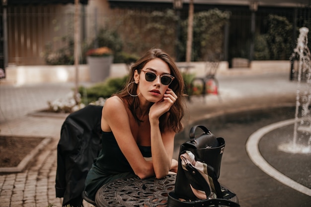 Bastante joven con peinado ondulado oscuro, labios rojos y gafas de sol negras, con vestido verde antideslizante posando en la terraza de un café al aire libre durante el día