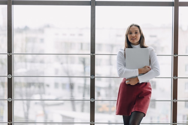 Bastante joven con el portátil junto a la ventana en la oficina con grandes ventanales. concepto de trabajo a distancia. independiente trabajando solo.