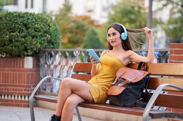 Bastante joven sentada en un banco en la ciudad escuchando música con auriculares mirando a otro lado