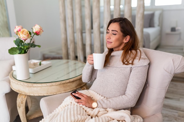 Bastante joven sentada en el sofá y disfrutando del primer café de la mañana en un acogedor apartamento. Mujer sonriente sentada en el sofá mientras bebe té caliente. Niña feliz descansando en casa en una brillante mañana de invierno.