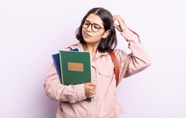 Bastante joven sintiéndose desconcertado y confundido, rascándose la cabeza. estudiante con concepto de libros