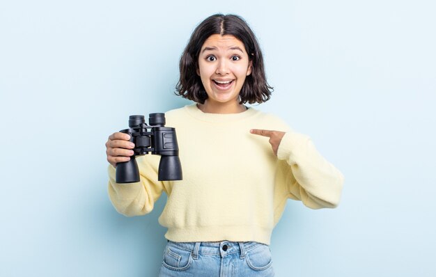 Bastante joven sintiéndose feliz y apuntando a sí mismo con un emocionado. concepto de binoculares