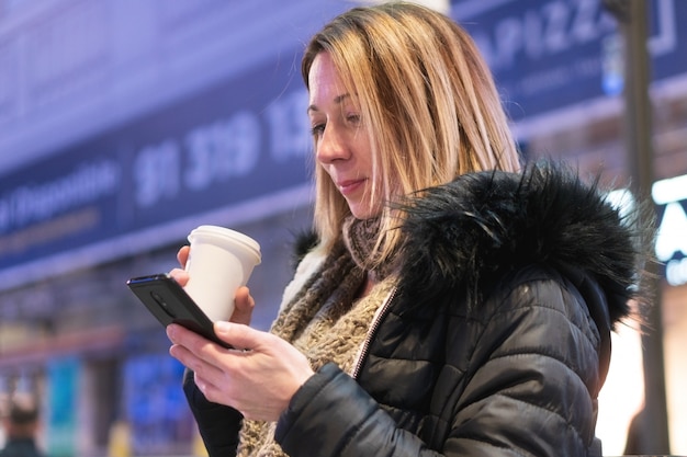 Bastante joven con smartphone y café sonriendo feliz en la ciudad por la noche.