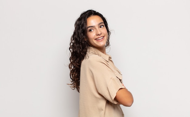 Bastante joven sonriendo alegremente, sintiéndose feliz, satisfecha y relajada, con los brazos cruzados y mirando hacia un lado