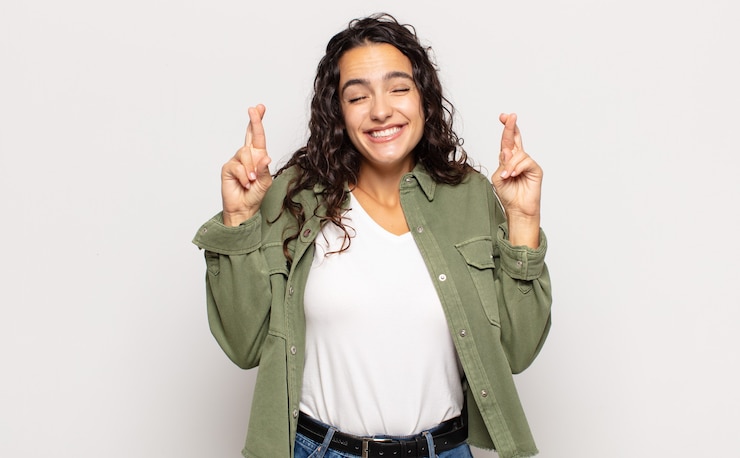 Foto bastante joven sonriendo y cruzando ansiosamente ambos dedos, sintiéndose preocupada y deseando o esperando buena suerte