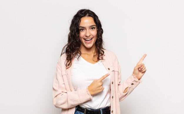 Bastante joven sonriendo felizmente y apuntando hacia un lado y hacia arriba con ambas manos mostrando el objeto en el espacio de la copia