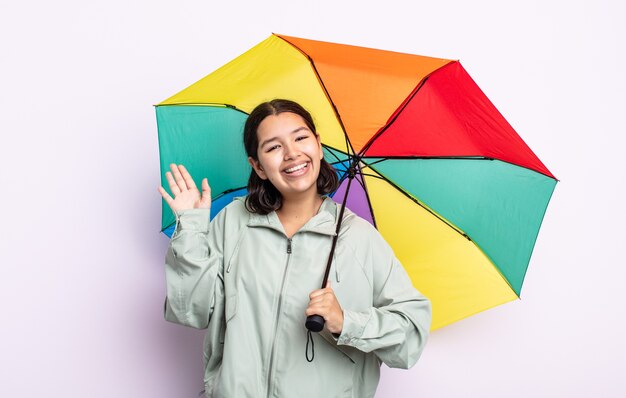 Bastante joven sonriendo felizmente, saludando con la mano, dándote la bienvenida y saludándote. concepto de lluvia y paraguas