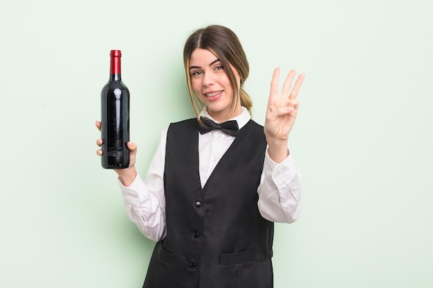 Bastante joven sonriendo y luciendo amigable, mostrando el número tres. concepto de camarero y botella de vino