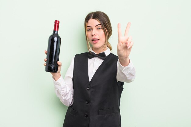 Bastante joven sonriendo y luciendo feliz gesticulando victoria o concepto de mesero de paz y botella de vino