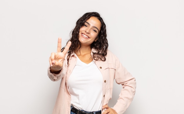 Bastante joven sonriendo y mirando amistosamente, mostrando el número dos o el segundo con la mano hacia adelante, contando hacia atrás