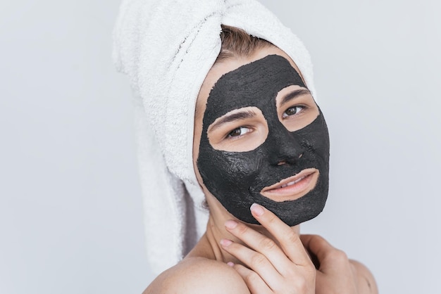 Bastante joven sonriente hermosa mujer aplicando máscara de arcilla negra haciendo tratamiento de bienestar de belleza en la piel de la cara Tiro horizontal de mujer aplicando máscara facial orgánica posando contra la pared blanca del estudio