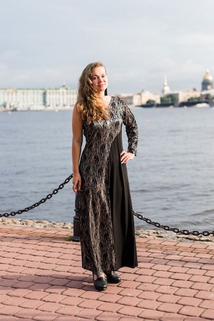 Bastante joven sonriente mujer caucásica en vestido gris y negro hasta el suelo en el muelle.