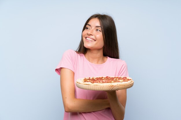 Foto bastante joven sosteniendo una pizza sobre la pared azul aislada mirando hacia arriba mientras sonríe