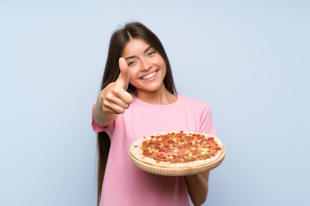 Bastante joven sosteniendo una pizza sobre la pared azul aislada con los pulgares hacia arriba porque algo bueno ha sucedido