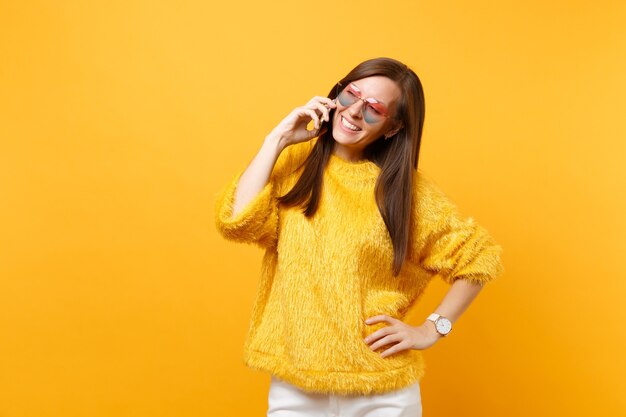 Bastante joven en suéter de piel, gafas de corazón hablando por teléfono móvil, llevando a cabo una agradable conversación aislada sobre fondo amarillo brillante. Personas sinceras emociones, estilo de vida. Área de publicidad.