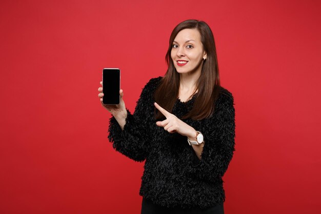 Bastante joven en suéter de piel negra apuntando con el dedo índice en el teléfono móvil con pantalla vacía en blanco aislada sobre fondo rojo brillante. Personas sinceras emociones, concepto de estilo de vida. Simulacros de espacio de copia.