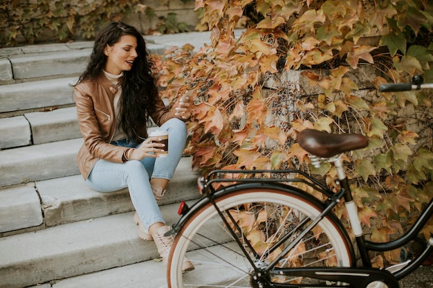 Bastante joven con teléfono móvil beber café para ir a las escaleras en bicicleta el día de otoño