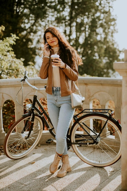 Bastante joven con teléfono móvil beber café para llevar en bicicleta el día de otoño