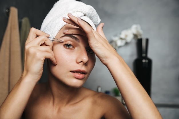 Bastante joven con una toalla en la cabeza corrigiendo las cejas con pinzas delante del espejo en el baño.