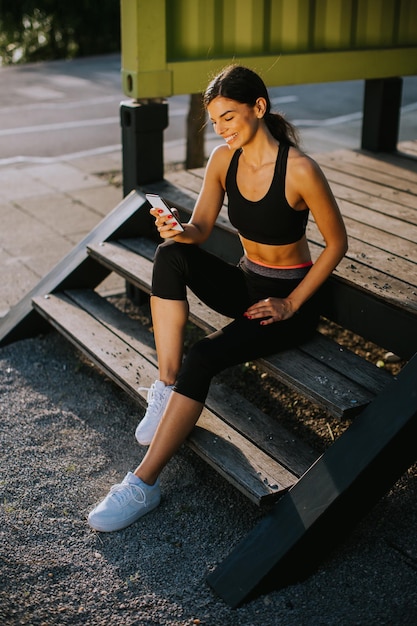 Bastante joven tomando un descanso durante el ejercicio al aire libre y usando el teléfono móvil