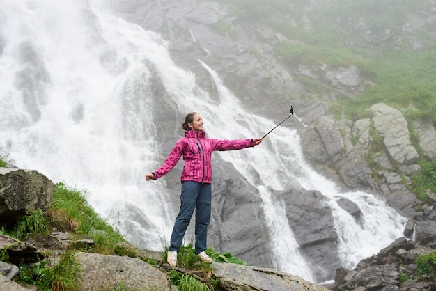 Bastante joven tomando un selfie frente a la gran cascada de gran alcance. Hermosa niña sonriente viajar en la naturaleza. Concepto de viaje y recreación.