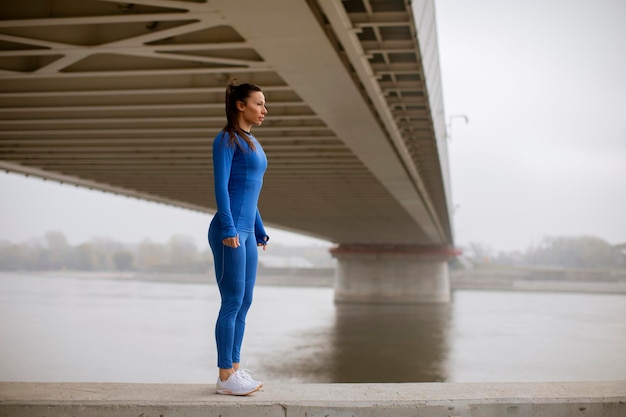 Foto bastante joven en traje azul que se extiende antes de entrenar por el río en la mañana de otoño