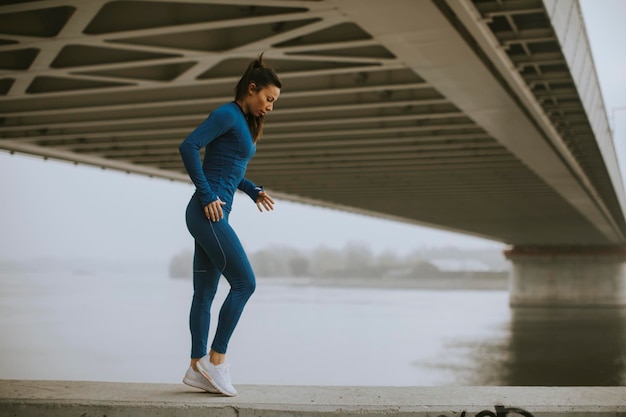 Bastante joven en traje azul que se extiende antes de entrenar por el río en la mañana de otoño