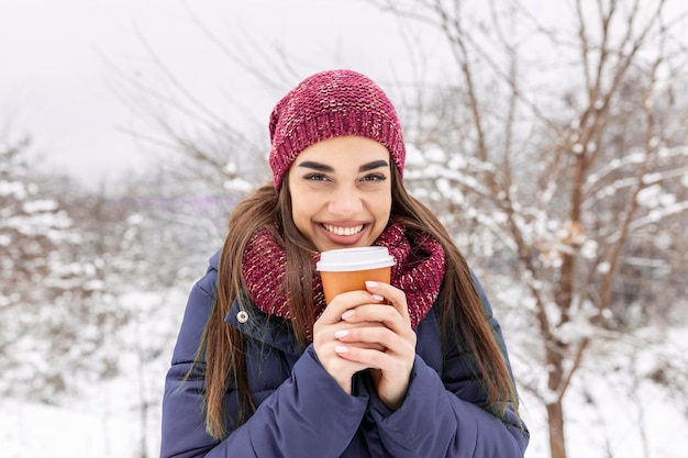 Bastante joven en traje de invierno sosteniendo una taza desechable llena de té o café caliente. Chica sosteniendo una taza de bebida caliente en sus manos y camina al aire libre en invierno.