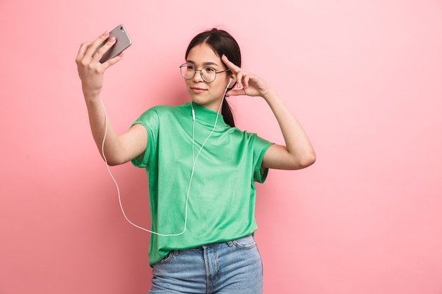 Bastante joven vistiendo anteojos redondos tomando selfie en teléfono móvil mientras usa auriculares aislados sobre pared rosa