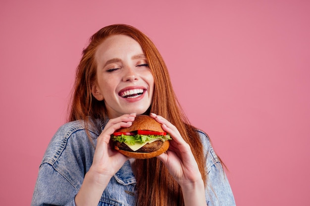 Bastante larga pelirroja jengibre chica ojos cerrados de placer comiendo papas fritas pollo y hamburguesa con papas fritas en fondo rosa estudio.