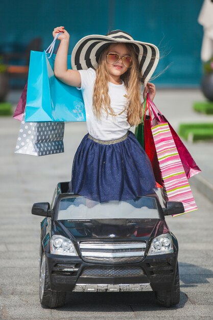 Bastante niña fashionista haciendo compras con bolsas de compras. Niño lindo al aire libre en el coche de juguete