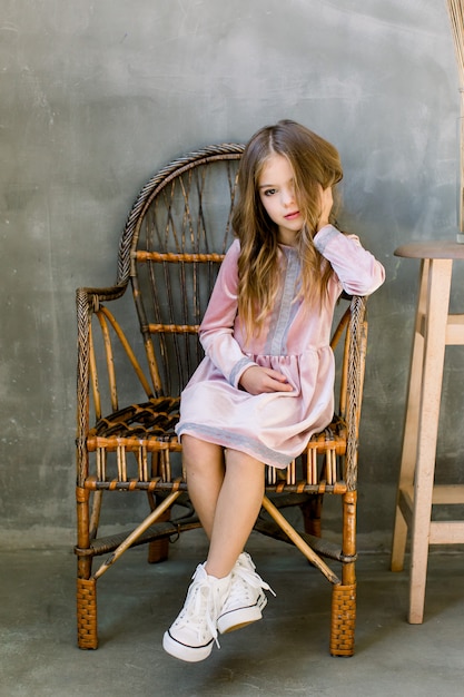 Bastante pequeña niña en un hermoso vestido rosa se sienta en la silla de madera y se ve soñadora, concepto de belleza y moda, retrato interior