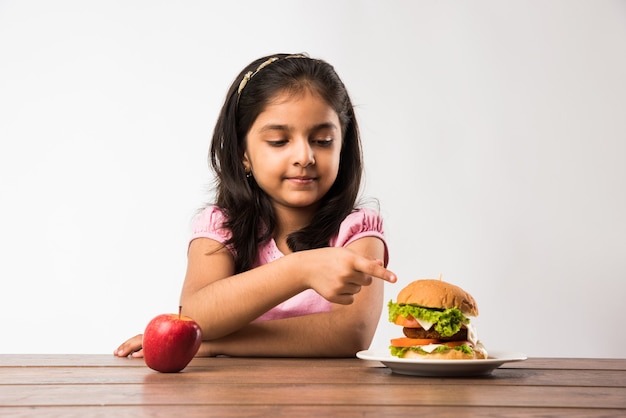 Bastante pequeña niña india o asiática incapaz de elegir entre manzana fresca y hamburguesa, concepto de alimentación saludable