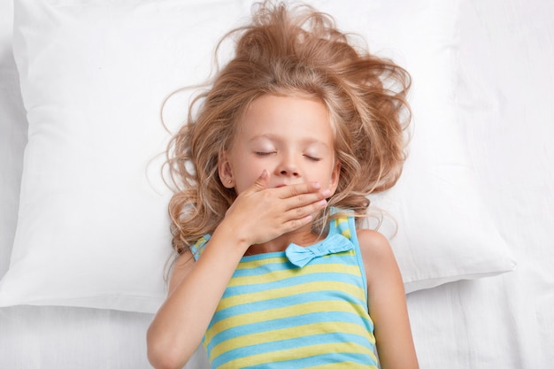 Bastante somnolienta hermosa niña pequeña mantiene los ojos cerrados, la palma de la mano en la boca, vestida con ropa de dormir casual a rayas, se acuesta en una cama cómoda, se despierta después de dormir. Niños, buenos días concepto