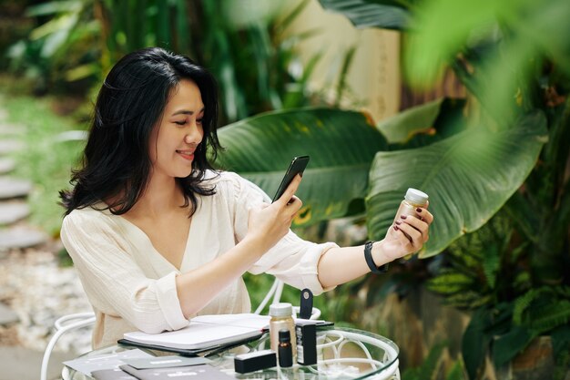 Bastante sonriente joven empresaria asiática tomando fotos de cosméticos para su tienda online