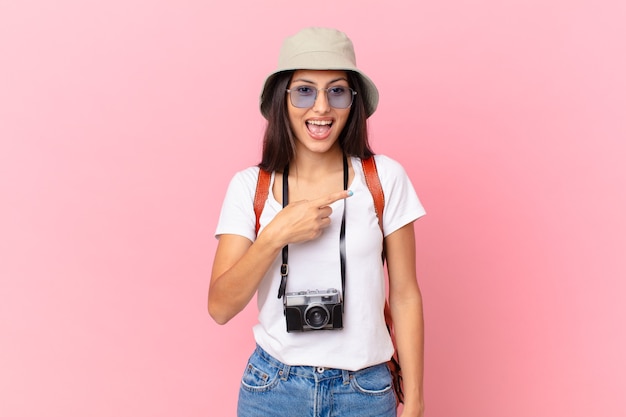 Foto bastante turista hispano mirando emocionado y sorprendido apuntando hacia el lado con una cámara de fotos y un sombrero