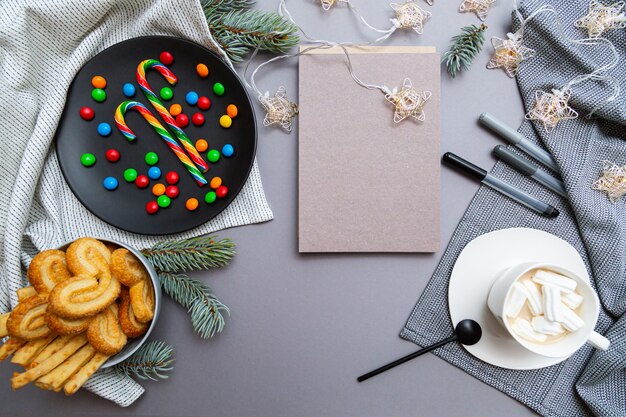 Bastones de caramelo de colores, galletas, guirnaldas de luces navideñas, cuaderno en blanco, taza de café y ramas de abeto