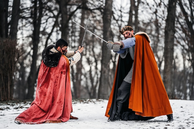 Batalha de dois cavaleiros em roupas e chapéus coloridos perto da floresta e da fortaleza de madeira. Cavaleiros lutam no inverno, na neve