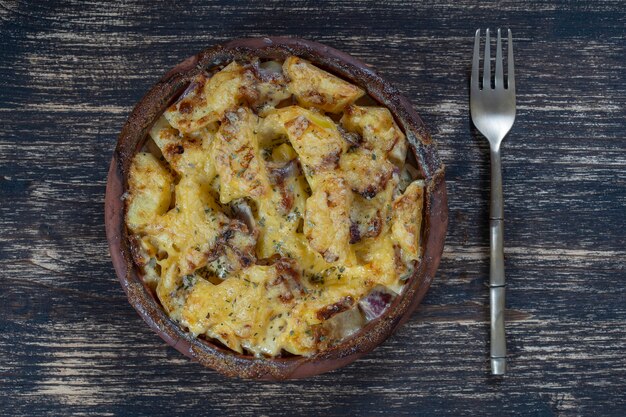 Batata assada, cebola e alho com queijo na mesa de madeira, close-up. Tigela de cerâmica com fundo de batata cebola e fatias de alho, vista superior