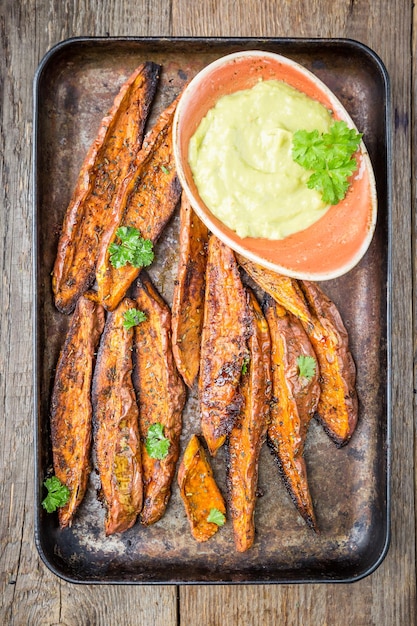 Batata doce assada caseira com ervas, sal e pimenta servida com molho de guacamole, em fundo de madeira, vista superior