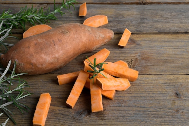 Batata-doce de laranja inteira crua e fatias de batatas com alecrim fresco em fundo de madeira rústico