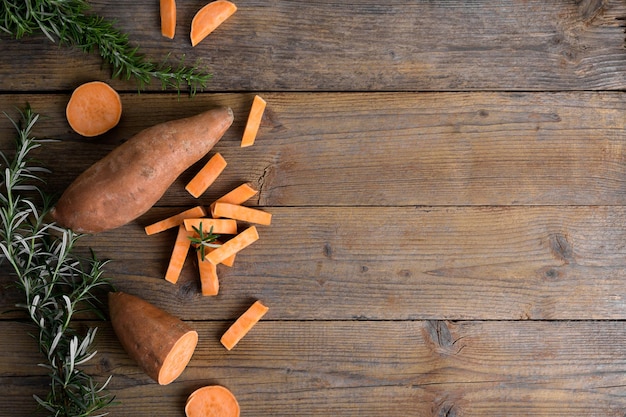 Batata naranja entera cruda y rodajas de batatas con romero fresco sobre fondo rústico de madera sobre el espacio de copia