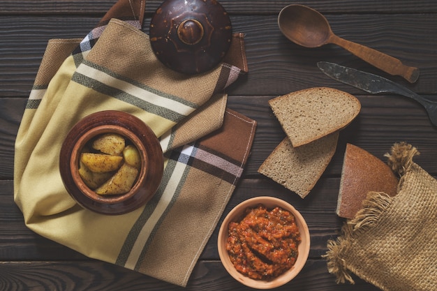 Batatas assadas em uma panela de barro, pão e molho sobre uma mesa de madeira