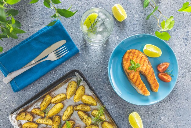 Foto batatas assadas na bandeja e bife de salmão grelhado com manjericão servido com gim-tônica com limão e alecrim na superfície da mesa cinza claro hora do jantar delicioso