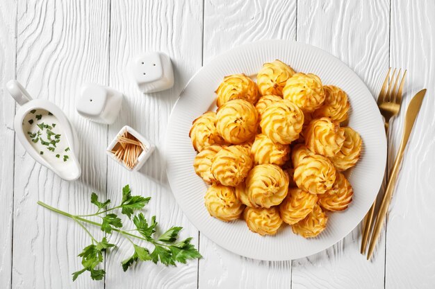 Batatas Duquesa, purê de batata temperado em espirais decorativos, douradas no forno e servidas com molho branco em um prato branco sobre uma mesa de madeira com talheres dourados