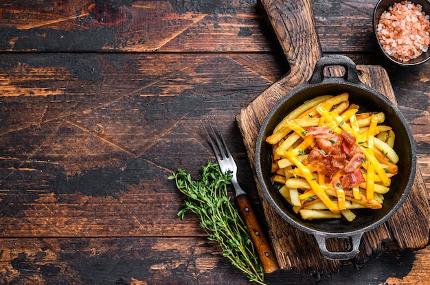 Batatas fritas com mistura derretida de queijo cheddar e bacon servidas na frigideira. Fundo de madeira escuro.